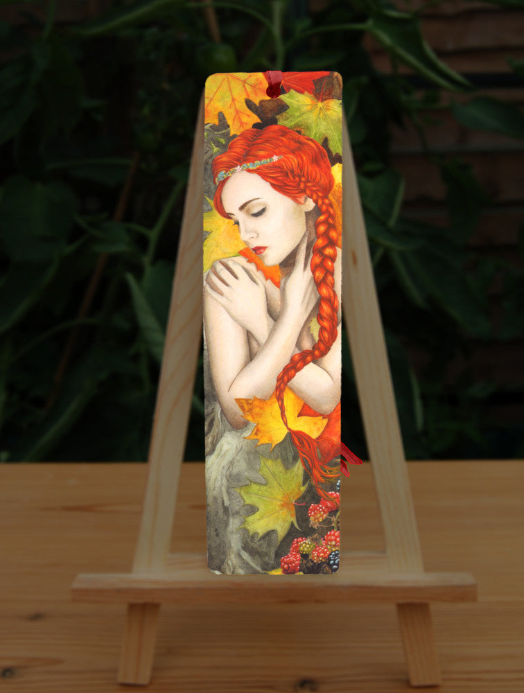 A woman with bright red hair, her bare arms folded across her chest with her hands over her shoulders. She is surrounded by large, autumn coloured leaves, and blackberries at the bottom.