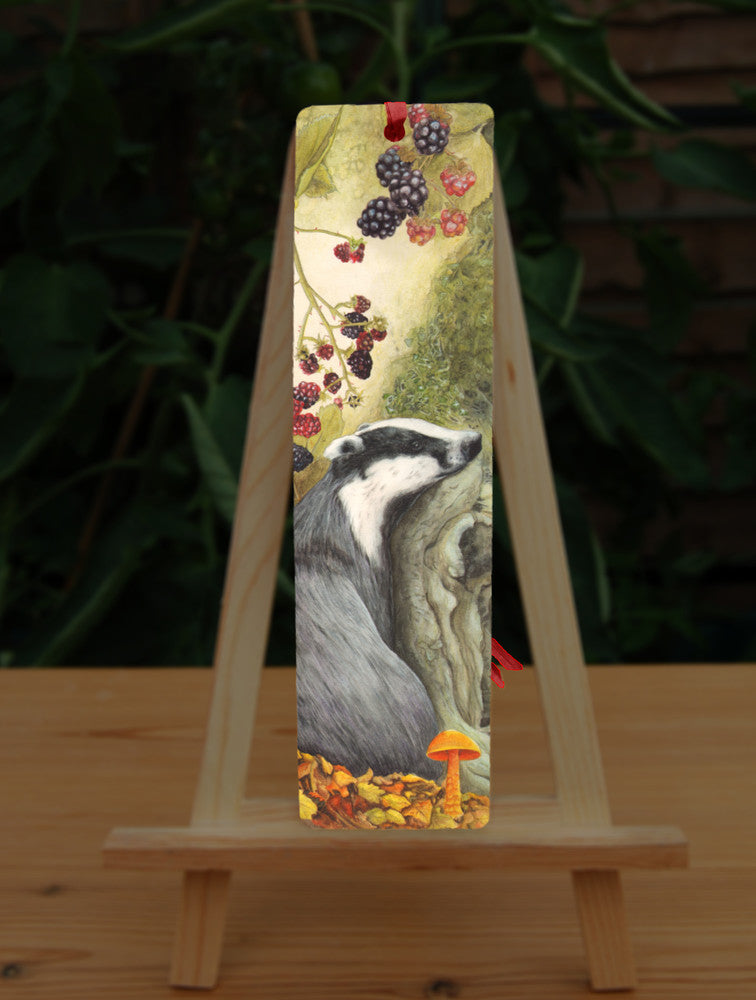A badger sits against a moss-covered tree trunk. Blackberries hang from above, with autumn leaves and mushrooms at its feet.