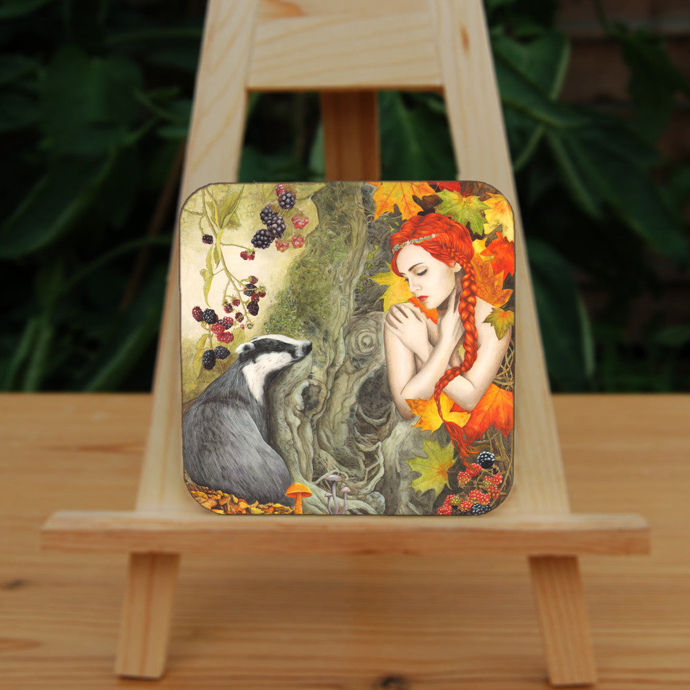 A woodland scene. On the left is a badger, sitting against a moss-covered tree trunk in the centre of the image, looking towards a crouching woman with bright red hair, her bare arms folded across her chest with her hands over her shoulders. She looks towards the badger with eyes part-closed. Blackberries hang above the badger, with autumn leaves and mushrooms at its feet. The woman is surrounded by large, autumn coloured leaves, with blackberries at her knees. Overall colours are oranges, reds and greens.