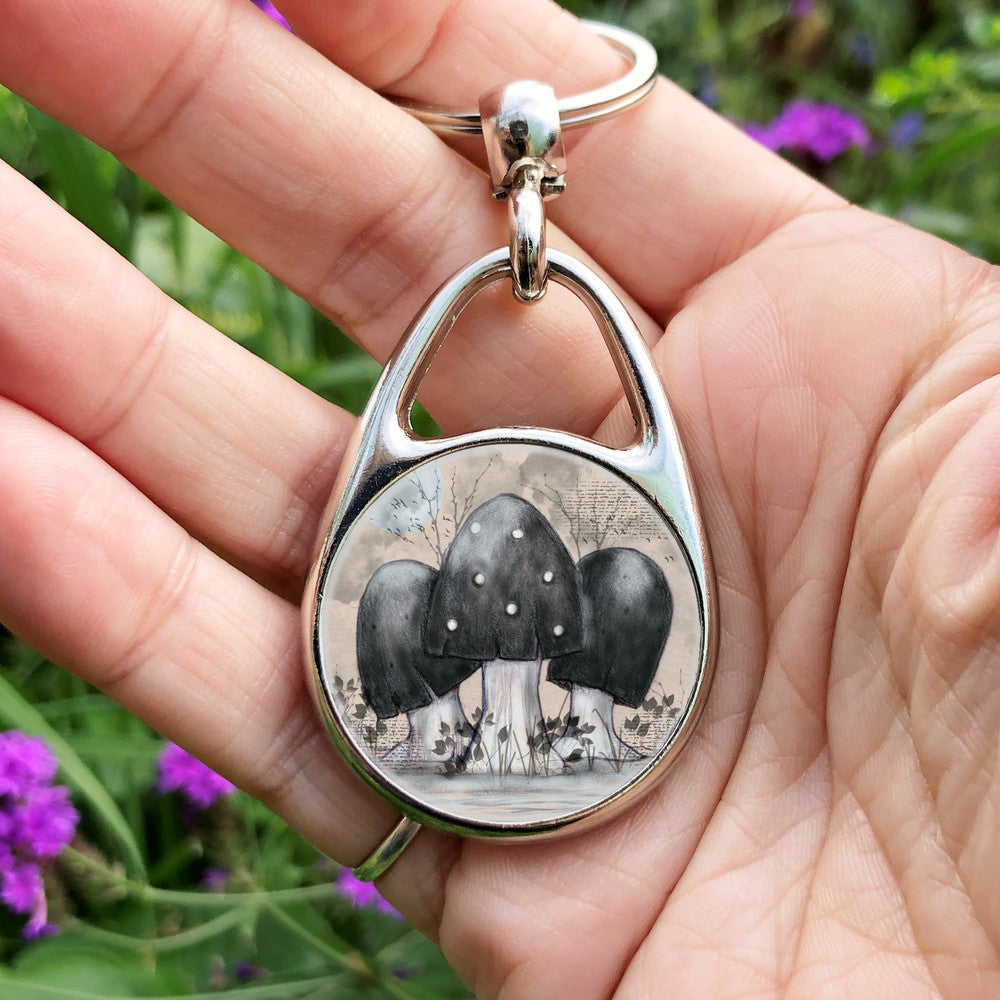 A cluster of toadstools with black caps. The cap of the centre toadstool has white spots. They're surrounded by the silhouettes of leafy plants, and a mist lies low on the ground. Above them a silvery full moon shines, and a flock of birds flies across. The background is a mottled beige.