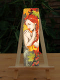 A woman with bright red hair, her bare arms folded across her chest with her hands over her shoulders. She is surrounded by large, autumn coloured leaves, and blackberries at the bottom.