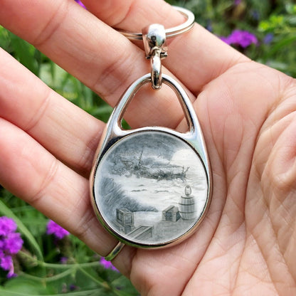 A blue-tinted pencil drawing. Scattered wooden crates and a wooden barrel with an oil lamp on top look down and across a large cave. The sea laps against the shore below, and on the other side of the cave, a little way in from the entrance, the remains of a ship lies broken and rotting.