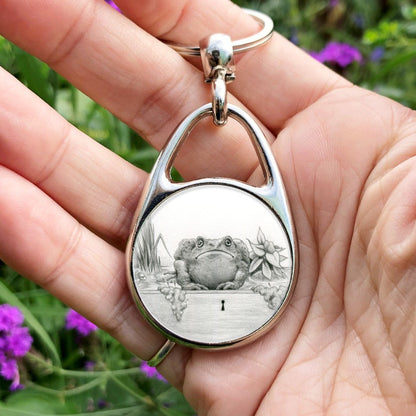 Black and white pencil drawing of fat toad sitting facing the viewer, front legs angled under its chest. It's sitting in a water-filled drawer (the keyhole can be seen at the front) filled with plants and flowers.