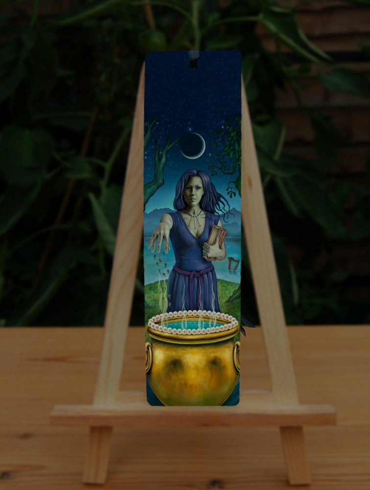 A woman with dark hair and wearing a blue dress with purple woven belt, and a necklace of three bones, stands behind a gold cauldron, her arm outstretched as she drops something into it. She looks towards the viewer. In her other arm she holds a book with '13' written on it. She stands between two trees, one an oak, the other unknown, on a small island of grass with water all around. Behind her, a harp floats in the water. A crescent moon rises above her. Mountains can be seen in the far background.