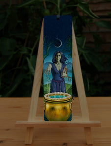 A woman with dark hair and wearing a blue dress with purple woven belt, and a necklace of three bones, stands behind a gold cauldron, her arm outstretched as she drops something into it. She looks towards the viewer. In her other arm she holds a book with '13' written on it. She stands between two trees, one an oak, the other unknown, on a small island of grass with water all around. Behind her, a harp floats in the water. A crescent moon rises above her. Mountains can be seen in the far background.
