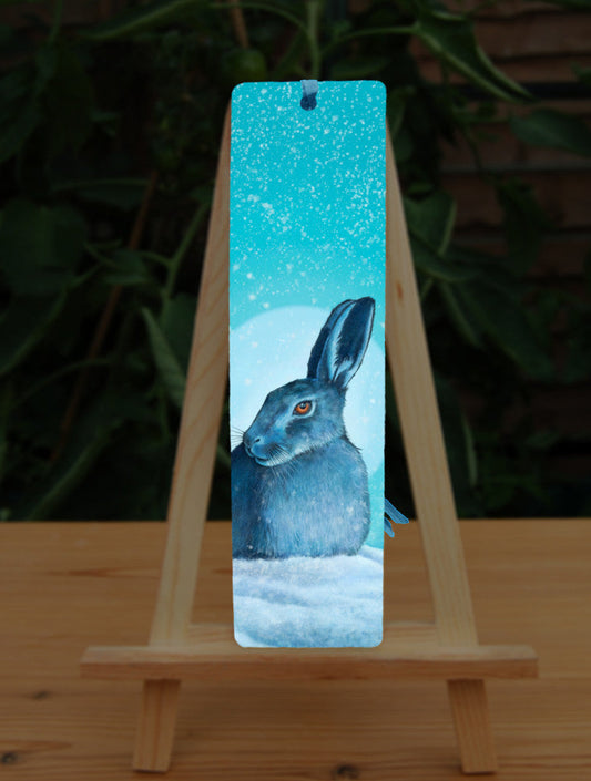 A hare crouches in snow, head turned to one side so one eye faces the viewer. Snow is falling, and a pale wintery sun shines behind. The design is coloured in shades of blue and turquoise, including the hare, which appears blue except for a deep orange eye.