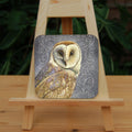 Square coaster: A pale brown bird with cream face sits sideways, coming in from the left hand side of the design, and looking directly at the viewer. The background is grey, overlaid a paler grey border consisting of a pattern of swirls.