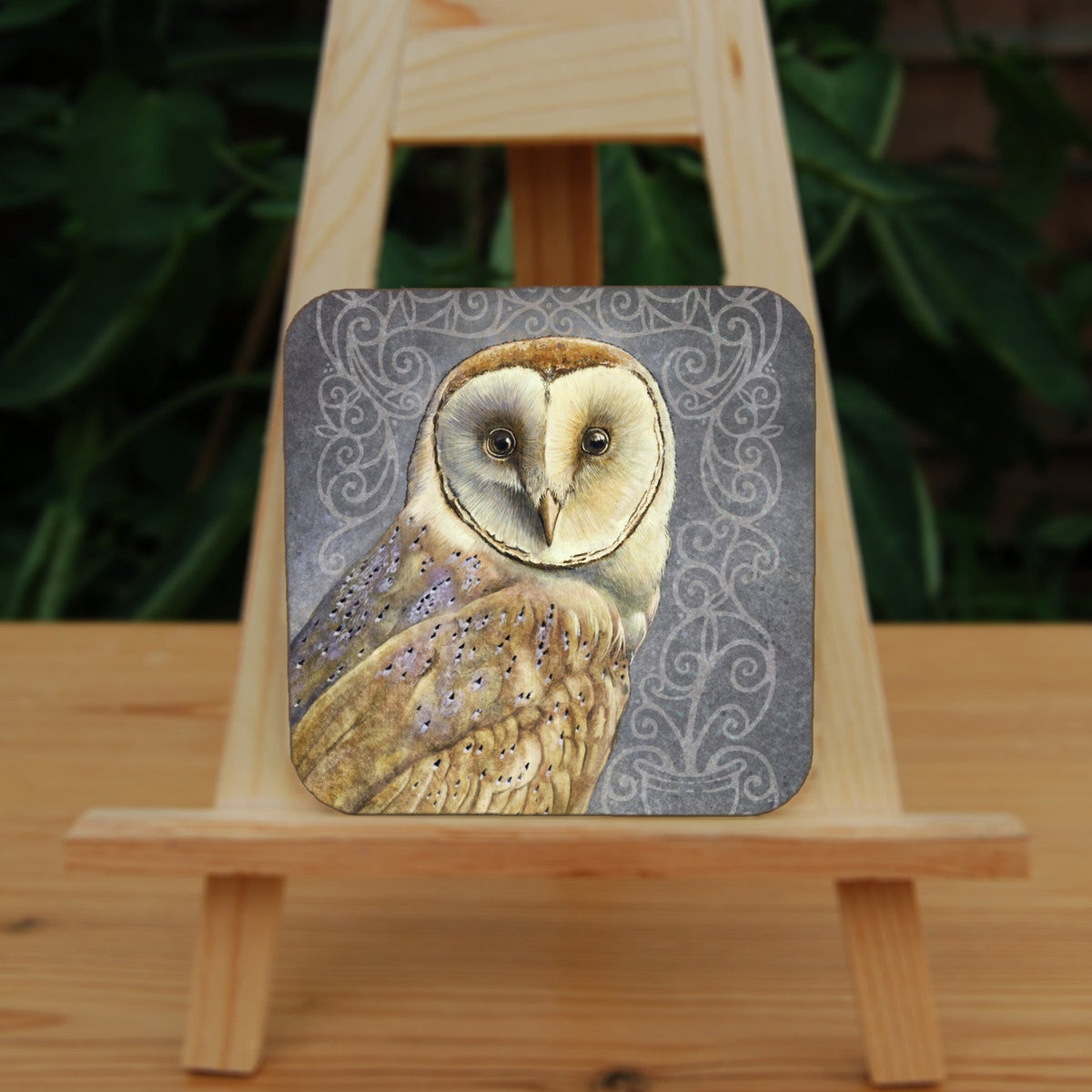 Square coaster: A pale brown bird with cream face sits sideways, coming in from the left hand side of the design, and looking directly at the viewer. The background is grey, overlaid a paler grey border consisting of a pattern of swirls.