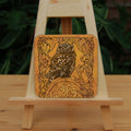 Square coaster: A compact-shaped owl with brown feathers sits sideways on an upright, roughly semi-circular stone arch, while looking directly at the viewer. The arch is decorated with a central circular Celtic knotwork design. A border of ivy leaves and simple curls surrounds the owl and down each side of the arch. The design is mostly monochrome apart from the owl, with a background of mottled dark tan.