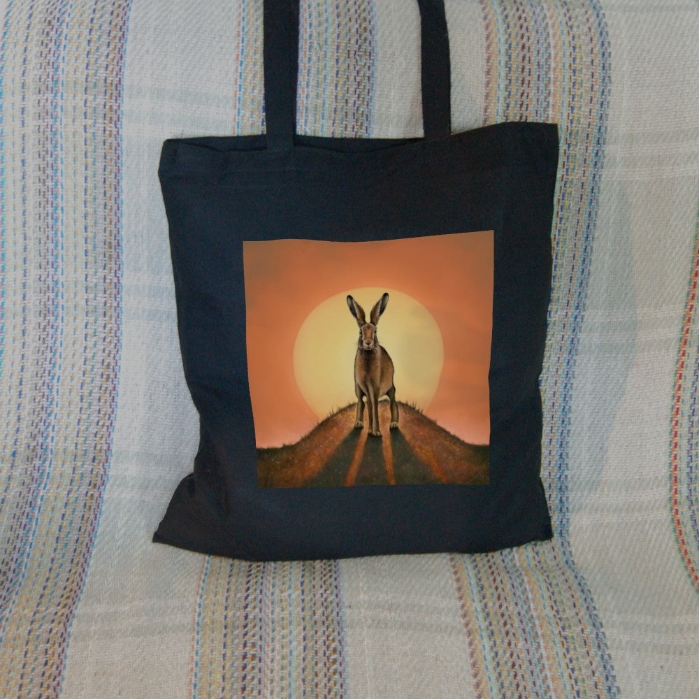 A brown hare stands on top of a small hill facing the viewer. The sun is rising behind the hare. The colours are mostly yellow, orange and brown.