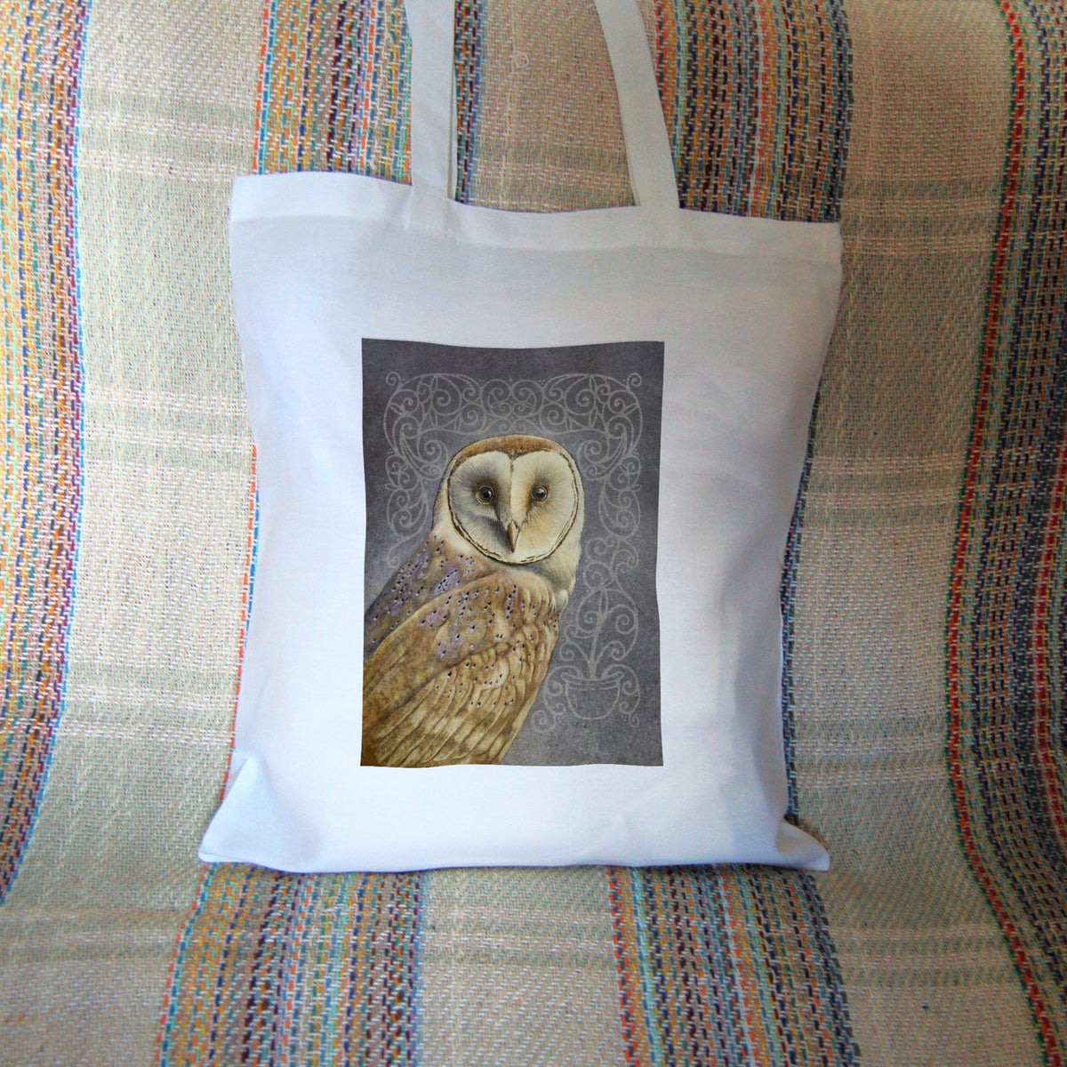 White cotton tote bag: A pale brown bird with cream face sits sideways, coming in from the left hand side of the design, and looking directly at the viewer. The background is grey, overlaid a paler grey border consisting of a pattern of swirls, ending with a pot at the bottom right from which the border appears to originate.