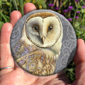 A hand holding a circular object: A pale brown bird with cream face sits sideways, coming in from the left hand side of the design, and looking directly at the viewer. The background is grey, overlaid a paler grey border consisting of a pattern of swirls.