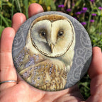 A hand holding a circular object: A pale brown bird with cream face sits sideways, coming in from the left hand side of the design, and looking directly at the viewer. The background is grey, overlaid a paler grey border consisting of a pattern of swirls.