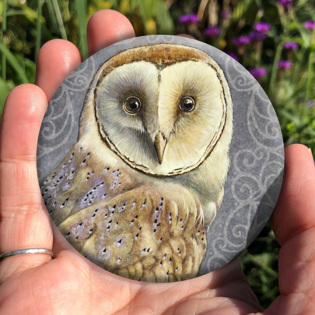 A hand holding a circular object: A pale brown bird with cream face sits sideways, coming in from the left hand side of the design, and looking directly at the viewer. The background is grey, overlaid a paler grey border consisting of a pattern of swirls.