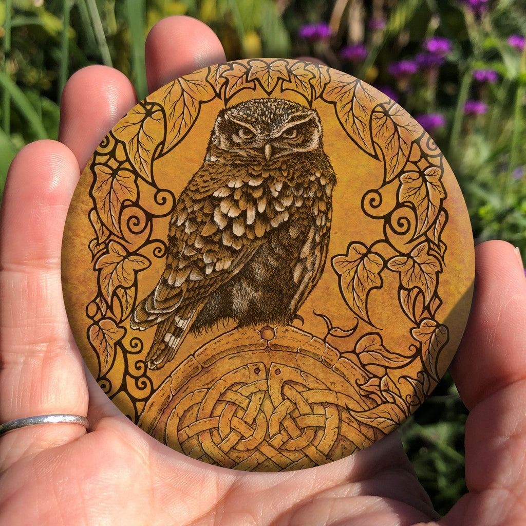 Hand holding a circular object: A compact-shaped owl with brown feathers sits sideways on an upright, roughly semi-circular stone arch, while looking directly at the viewer. The arch is decorated with a central circular Celtic knotwork design. A border of ivy leaves and simple curls surrounds the owl and down each side of the arch. The design is mostly monochrome apart from the owl, with a background of mottled dark tan.