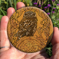 Hand holding a circular object: A compact-shaped owl with brown feathers sits sideways on an upright, roughly semi-circular stone arch, while looking directly at the viewer. The arch is decorated with a central circular Celtic knotwork design. A border of ivy leaves and simple curls surrounds the owl and down each side of the arch. The design is mostly monochrome apart from the owl, with a background of mottled dark tan.