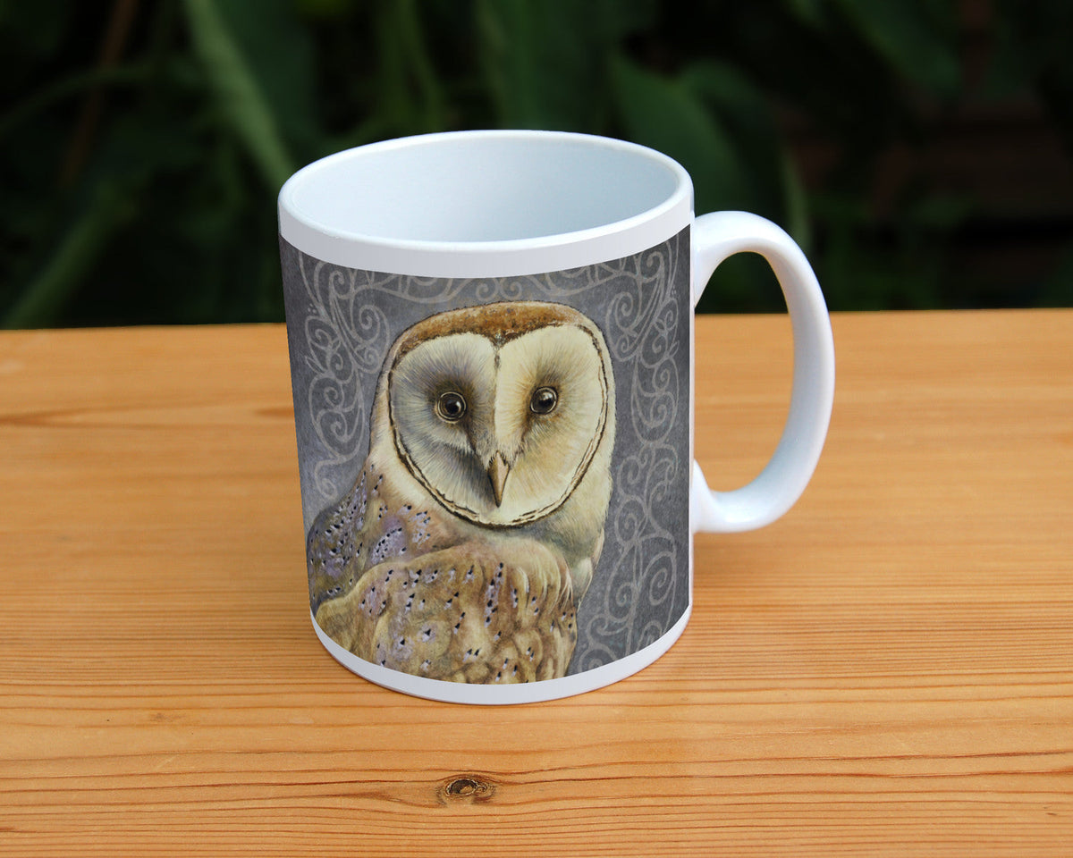 A white ceramic mug: A pale brown bird with cream face sits sideways, coming in from the left hand side of the design, and looking directly at the viewer. The background is grey, overlaid a paler grey border consisting of a pattern of swirls.