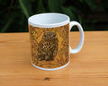 A white ceramic mug: A compact-shaped owl with brown feathers sits sideways on an upright, roughly semi-circular stone arch, while looking directly at the viewer. A border of ivy leaves and simple curls surrounds the owl and down each side of the arch. The design is mostly monochrome apart from the owl, with a background of mottled dark tan.