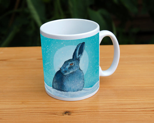 A hare crouches in snow, head turned to one side so one eye faces the viewer. Snow is falling, and a pale wintery sun shines behind. The design is coloured in shades of blue and turquoise, including the hare, which appears blue except for a deep orange eye.