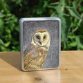 A rectangular metal box: A pale brown bird with cream face sits sideways, coming in from the left hand side of the design, and looking directly at the viewer. The background is grey, overlaid a paler grey border consisting of a pattern of swirls, ending with a pot at the bottom right from which the border appears to originate.