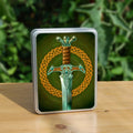 A shining silver sword stood upright, the handle and part of the blade visible. The handle is leather, and there's a face in the pommel. A bronze-coloured Celtic knot circle sits behind the sword. The background is green.