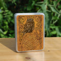A rectangular metal tin: A compact-shaped owl with brown feathers sits sideways on an upright, roughly semi-circular stone arch, while looking directly at the viewer. The arch is decorated with a central circular Celtic knotwork design. A border of ivy leaves and simple curls surrounds the owl and down each side of the arch. The design is mostly monochrome apart from the owl, with a background of mottled dark tan.