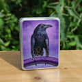 A raven sitting on a branch facing the viewer with their head turned sideways and wings slightly open. The raven is coloured black with purple shading, and the background is mottled purple.