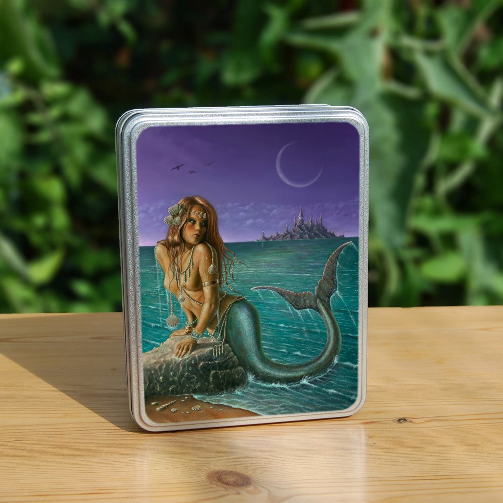 A mermaid leans on a rock, using her arms to hold herself clear of the water. She's on the shoreline, and is looking over her shoulder, across the teal-coloured sea, towards a rocky island with tall, pointed buildings on top. The sky is purple, and a crescent moon and flying seabirds are visible. The mermaid is naked with a teal fish tail, curled up towards her back. There's silver jewellery draped around her body, and she has seashells in her long hair.