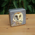 A square metal tin: A pale brown bird with cream face sits sideways, coming in from the left hand side of the design, and looking directly at the viewer. The background is grey, overlaid a paler grey border consisting of a pattern of swirls.