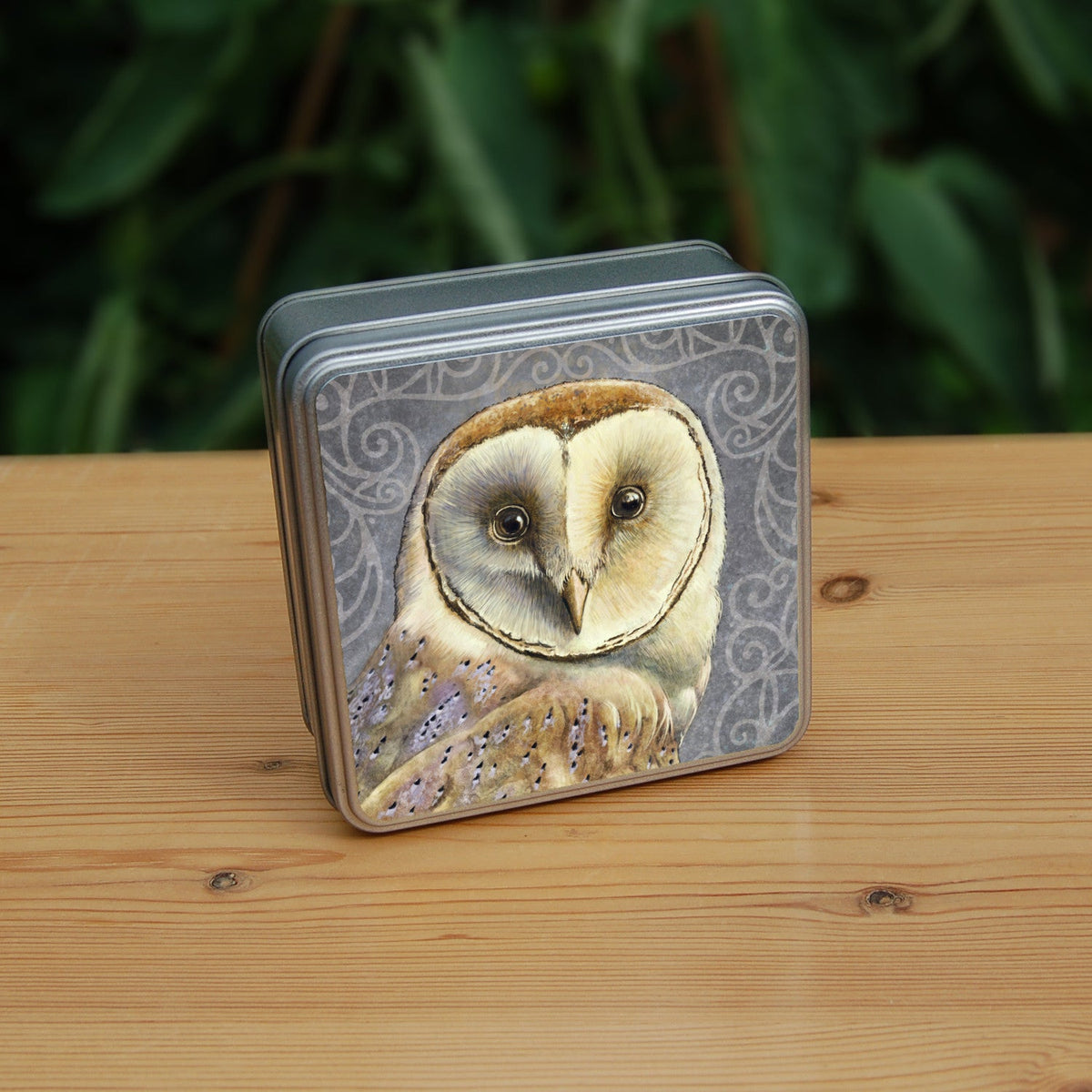 A square metal tin: A pale brown bird with cream face sits sideways, coming in from the left hand side of the design, and looking directly at the viewer. The background is grey, overlaid a paler grey border consisting of a pattern of swirls.