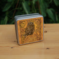 Square metal tin: A compact-shaped owl with brown feathers sits sideways on an upright, roughly semi-circular stone arch, while looking directly at the viewer. The arch is decorated with a central circular Celtic knotwork design. A border of ivy leaves and simple curls surrounds the owl and down each side of the arch. The design is mostly monochrome apart from the owl, with a background of mottled dark tan.