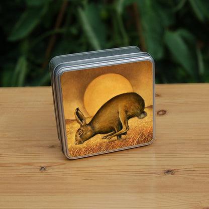 A brown hare runs from right to left, back curved, hind legs ahead of front legs. The sun rises behind, casting a golden brown glow across the sky, field and grass beneath the hare.