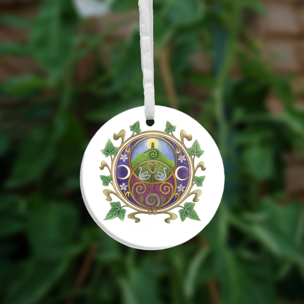 A circular design on a white background featuring the Chalice Well (Vesica Pisces) symbol inside a circle of ivy leaves. Glastonbury Tor, the sun rising behind the tower against a blue sky, nestles inside the upper circle of the symbol, with two swans in the area where the two circles join. An ammonite is inside the lower circle, against a maroon background. To the left and right are two opposing crescent moons, and small white flowers.