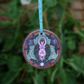 A raven stands behind a purple Goddess figure, its huge wings folded down her sides. She stands in the centre of a ring. Ravens fly around the inside of the ring, with each aspect of the triple moon also present. Colours are purple, red, and blue.