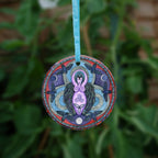 A raven stands behind a purple Goddess figure, its huge wings folded down her sides. She stands in the centre of a ring. Ravens fly around the inside of the ring, with each aspect of the triple moon also present. Colours are purple, red, and blue.