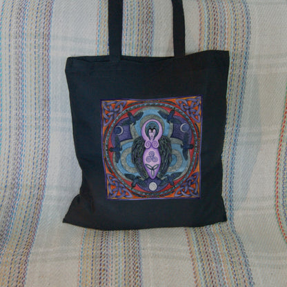 A raven stands behind a purple Goddess figure, its huge wings folded down her sides. She stands in the centre of a ring. Ravens fly around the inside of the ring, with each aspect of the triple moon also present. There's a Celtic knot in each corner. Colours are purple, red, and blue.