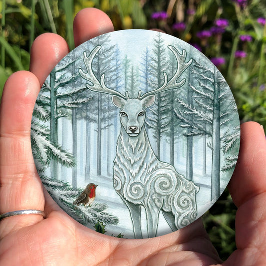 A blue-white stag with large swirls in his coat stands in snow facing the viewer. Snow-covered fir trees spread out behind him. A branch of berried holly can be seen on the bottom left, with a robin sitting on a fir tree branch above it.