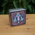 A raven stands behind a purple Goddess figure, its huge wings folded down her sides. She stands in the centre of a ring. Ravens fly around the inside of the ring, with each aspect of the triple moon also present. There's a Celtic knot in each corner. Colours are purple, red, and blue.
