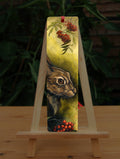 A rectangular bookmark: The head of a hare facing to the right. Sprigs of red rowan berries and leaves are drawn above and below. The background is mottled yellows and browns.