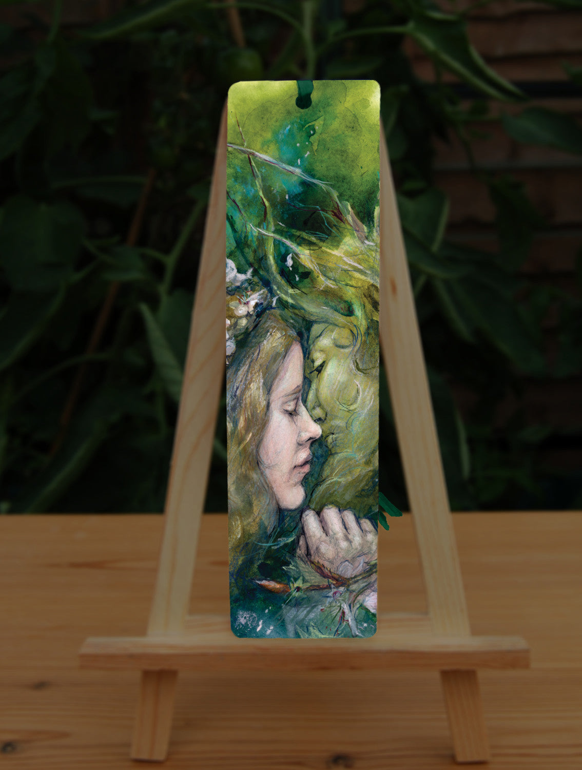 A rectangular bookmark: A young woman with a garland of pale flowers in her hair closes her eyes as she moves to kiss a green man.