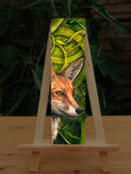 A tall bookmark: The head and neck of a red fox, their face angled towards the front right. The background consists of swirls of branches and leaves in a rich green colour.