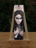 A tall bookmark: The head and hands of a woman. She has long dark hair and her face is decorated with patterns in blue paint. Her hands are held together against her body. On her wrist she wears a blue band with a raven skull attached. The background is a mottled brown.