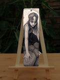 A tall bookmark: A young woman crouches on a pile of stones, her hands resting on stones in front of her. The woman wears a headband with a raven skull attached to the front. A full moon rises behind her.