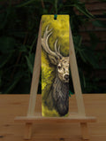 A rectangular bookmark: An off-to-one-side view of the head and chest of an antlered stag directly facing the viewer. Oak leaves are drawn to his left. The background is mottled yellows and browns.