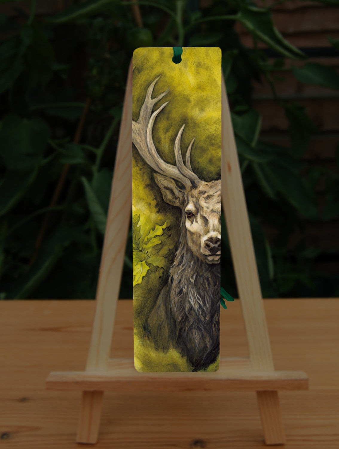 A rectangular bookmark: An off-to-one-side view of the head and chest of an antlered stag directly facing the viewer. Oak leaves are drawn to his left. The background is mottled yellows and browns.