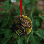 Circular hanging ornament with a red ribbon: The head and shoulders of a hare facing to the right. Sprigs of red rowan berries and leaves are drawn below. The background is mottled yellows and browns.