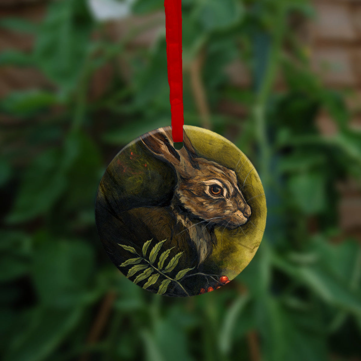 Circular hanging ornament with a red ribbon: The head and shoulders of a hare facing to the right. Sprigs of red rowan berries and leaves are drawn below. The background is mottled yellows and browns.