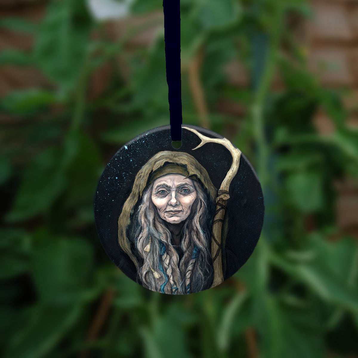 A circular item hanging against a leafy background: An older woman with blue eyes stands facing the viewer. Her head is covered by a brown hood, and her long grey hair, some of it braided with blue braid, falls down below her face. She holds a staff, topped with a deer antler, upright in one hand. A sea of stars shine behind her.