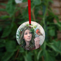 Circular hanging ornament with a red ribbon: A young woman looks towards a robin sitting at eye level on the fingers of her hand. She wears a red cloak with white fur trim, closed with a large Celtic brooch. Sprigs of red berries and ivy are woven into her hair. 