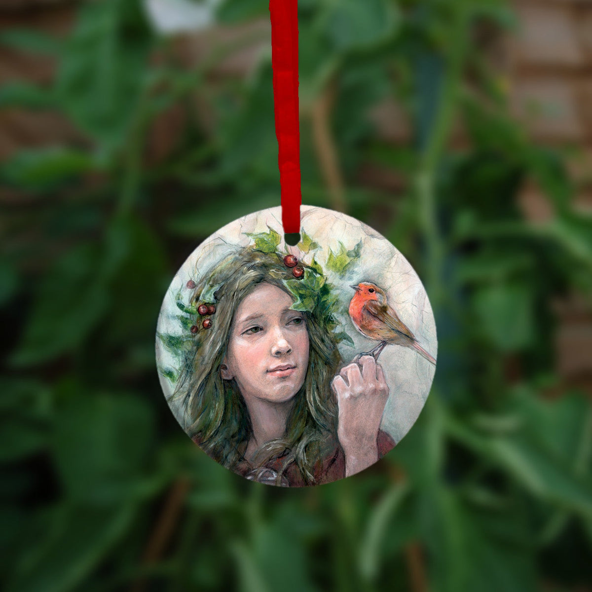 Circular hanging ornament with a red ribbon: A young woman looks towards a robin sitting at eye level on the fingers of her hand. She wears a red cloak with white fur trim, closed with a large Celtic brooch. Sprigs of red berries and ivy are woven into her hair. 